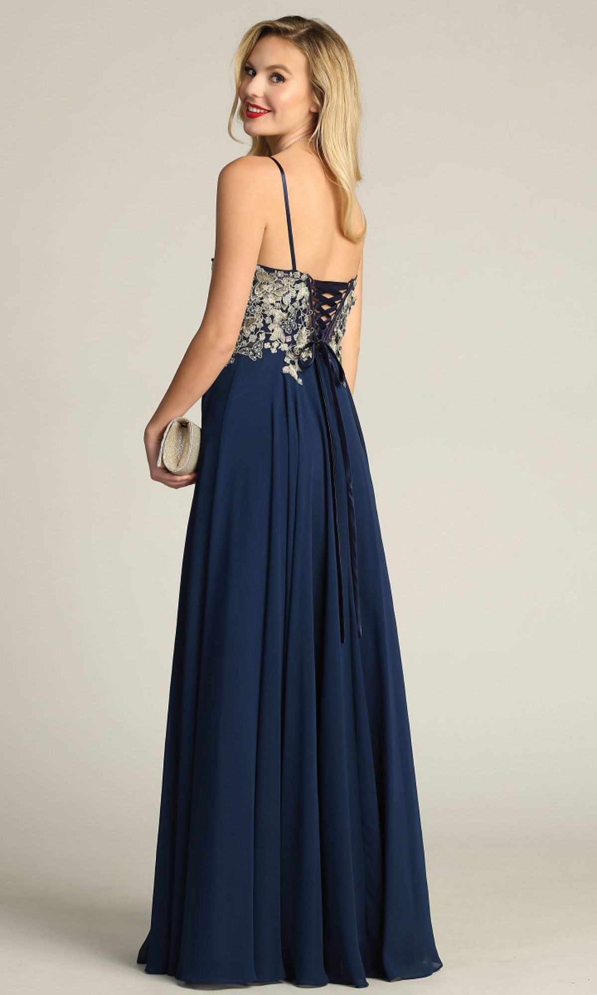 A woman in the Lace-Up Open-Back Long Prom Dress: Chicas C5124, a navy blue gown with thin straps and silver floral embroidery on the back, stands holding a clutch, facing away but glancing over her shoulder.